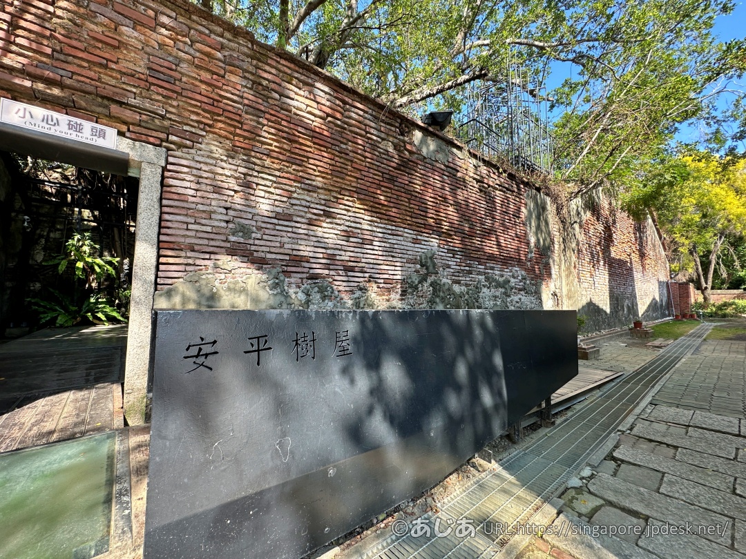 安平樹屋の黒い看板と、ガジュマルの木が覆いかぶさるレンガの壁