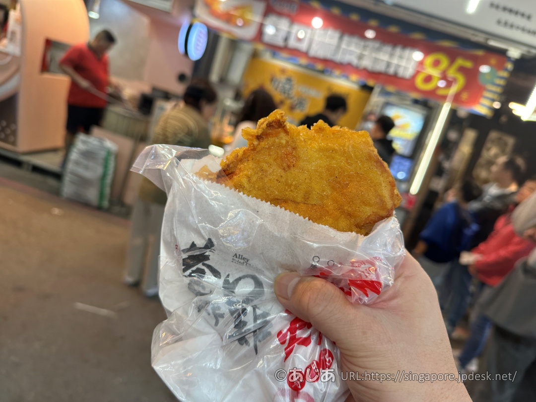 巷口炸雞のロゴが入ったビニール袋と紙袋に入れられた、黄金色の大きな特大フライドチキン(鶏排)を手で持っている様子