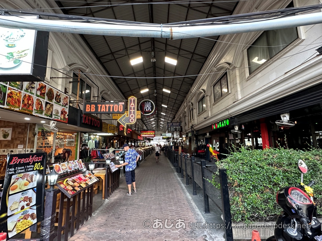 屋根付きのアーケード街の様子。両サイドにレストランやテーラー、タトゥーショップなどが並び、中央の通路を歩く人々の姿が見える。