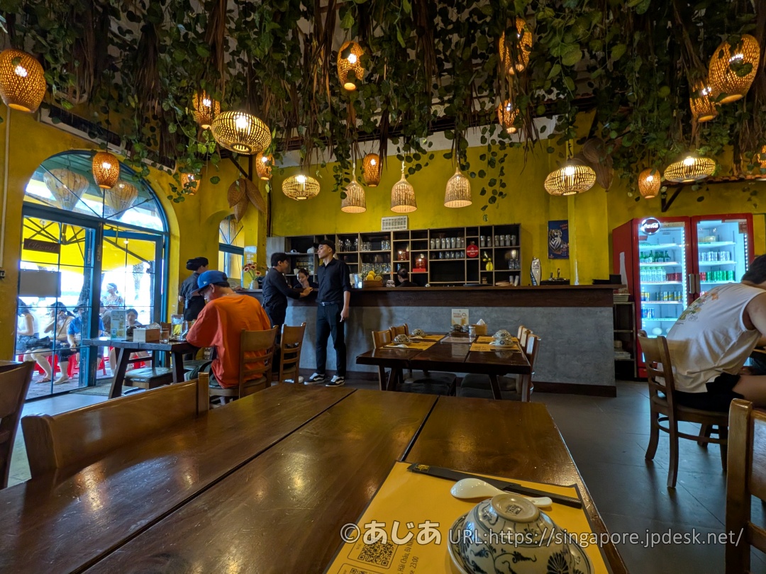 Ăn Thôiの店内。黄色い壁に、天井から竹編みのランプシェードと植物が吊るされている。木製のテーブルと椅子が並び、奥にはカウンターがある。