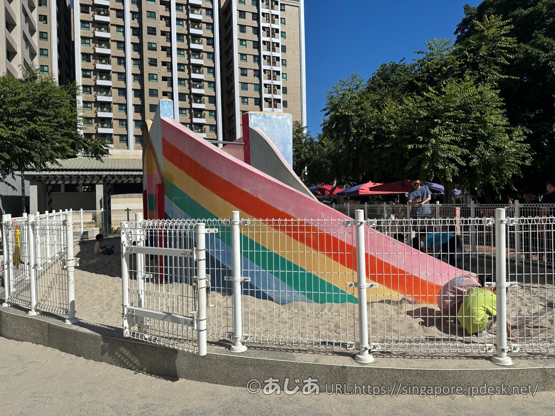 白い柵で囲まれた砂場にある大きなレインボーカラーの滑り台と、そこで遊ぶ子どもたちの様子。背景には緑の木々と近代的な高層マンションがそびえ立っている。