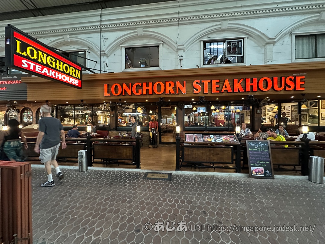 ロングホーン ステーキハウスの店頭。木目調の外観に赤と黄色のネオンサインで店名が掲げられており、テラス席にはお客さんが座っている。