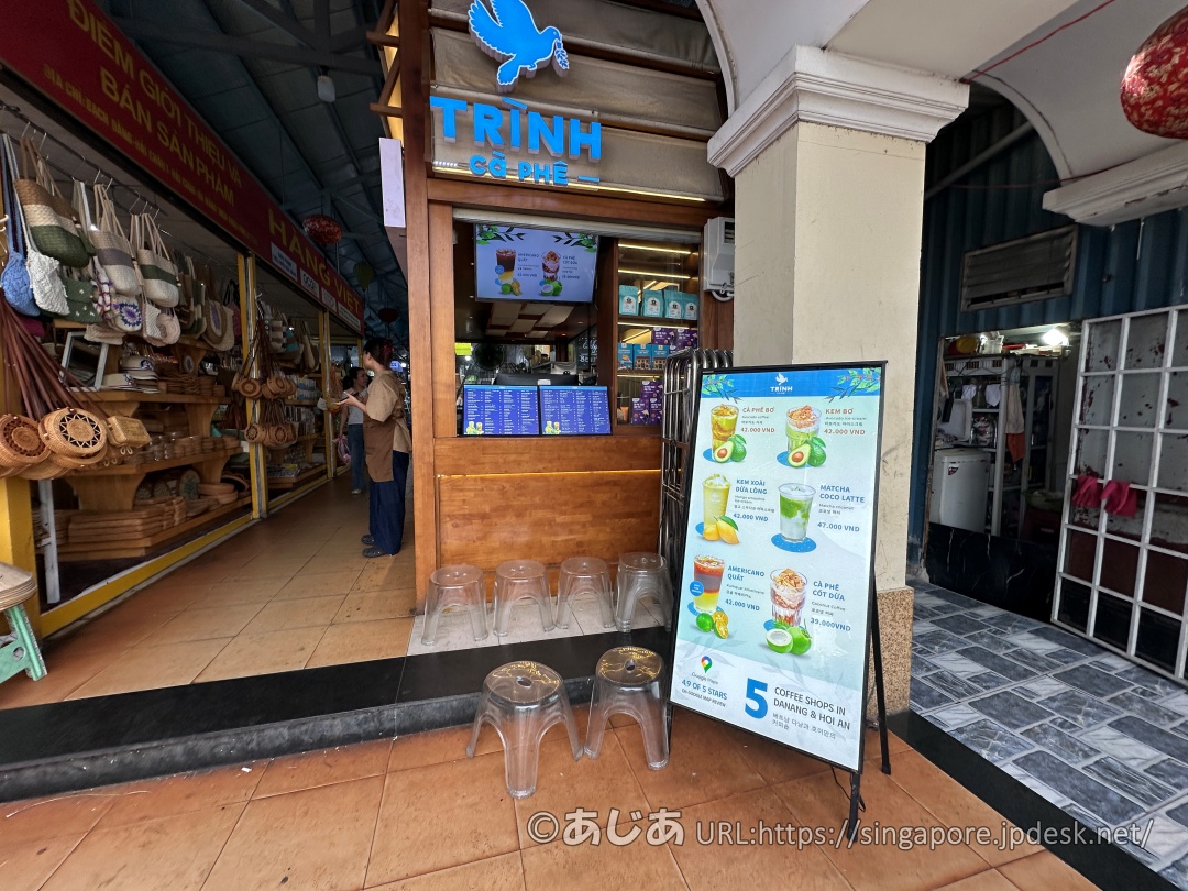 「TRỊNH CÀ PHÊ」の店舗正面。木目調のカウンターの前に、ドリンクの写真が載った立て看板が置かれている。手前には透明なプラスチックの小さな椅子が並んでいる。