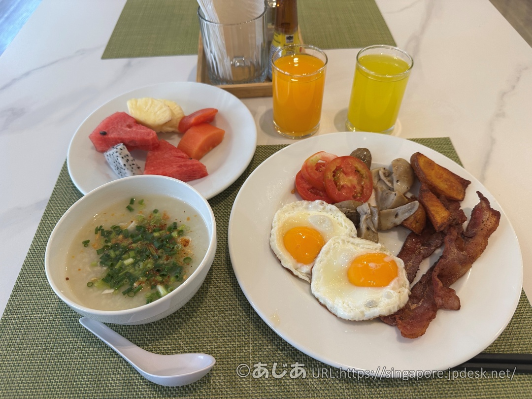 テーブルの上に並べられた朝食。目玉焼きやベーコンのお皿、ネギが乗ったお粥のボウル、フルーツのお皿、2種類のジュース。