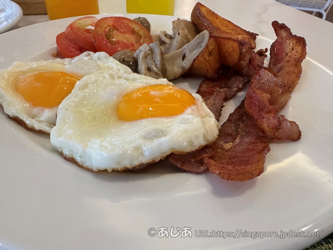 お皿に盛られた2つの目玉焼き、カリカリのベーコン、ポテト、マッシュルーム、トマトのアップ写真。