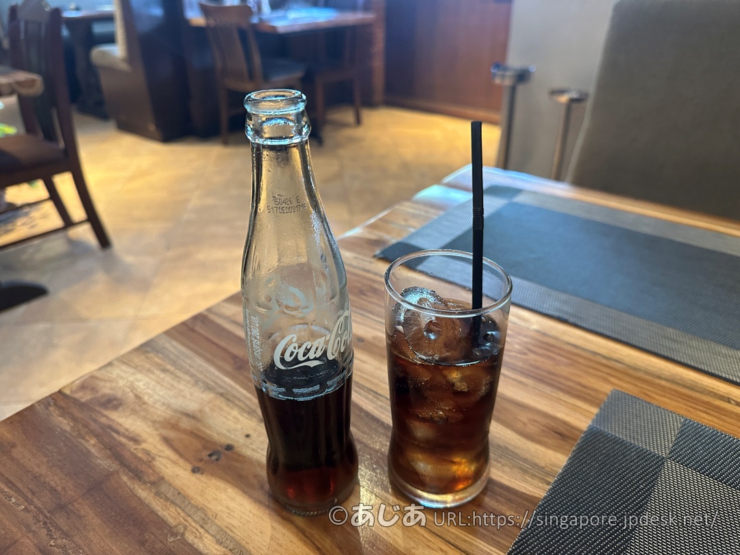 木製のテーブルの上に置かれた瓶入りのコカ・コーラと、氷が入ったグラス。