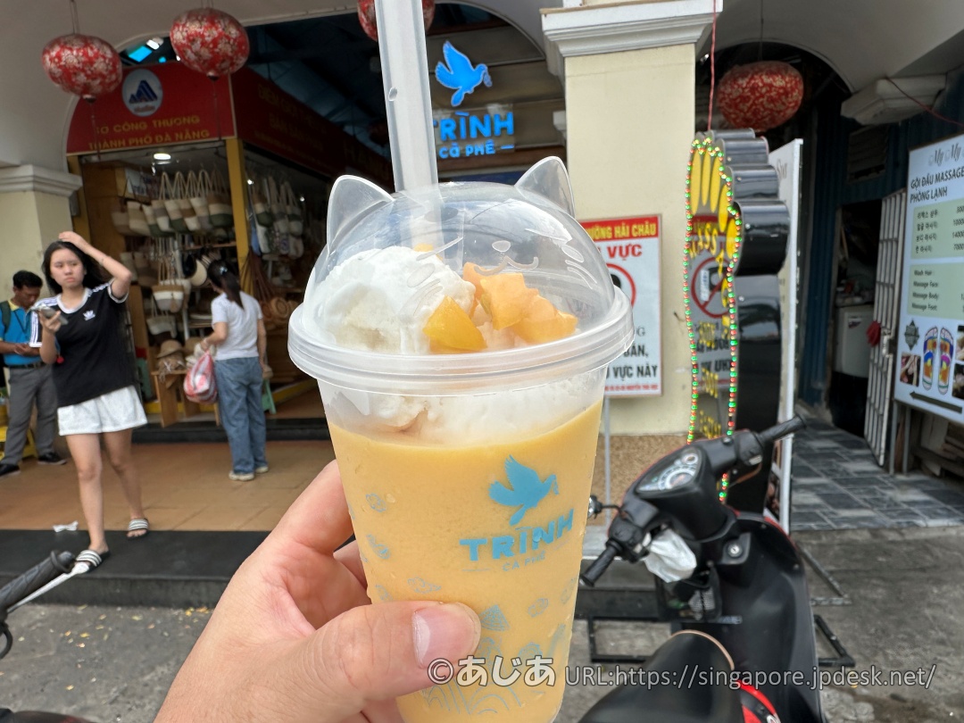 手に持ったマンゴードリンクのアップ。透明なカップには青い鳥のロゴがあり、フタには猫の耳がついている。中にはオレンジ色のマンゴーシェイク、白いアイスクリーム、角切りのマンゴー果肉が入っている。背景にはお店と通りが見える。