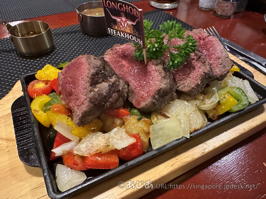 熱々の鉄板に乗った分厚いテンダーロインステーキ。下にはパプリカや玉ねぎなどのロースト野菜が敷き詰められ、お肉には店のロゴが入った小さな旗が刺さっている。