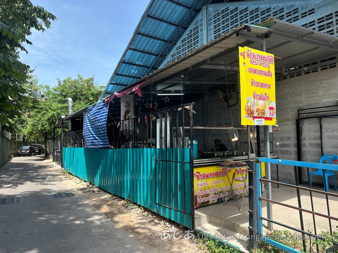 青いトタン屋根と緑色の囲いがある半屋外のタイ食堂の外観。入り口には写真付きの黄色い看板が立っている。