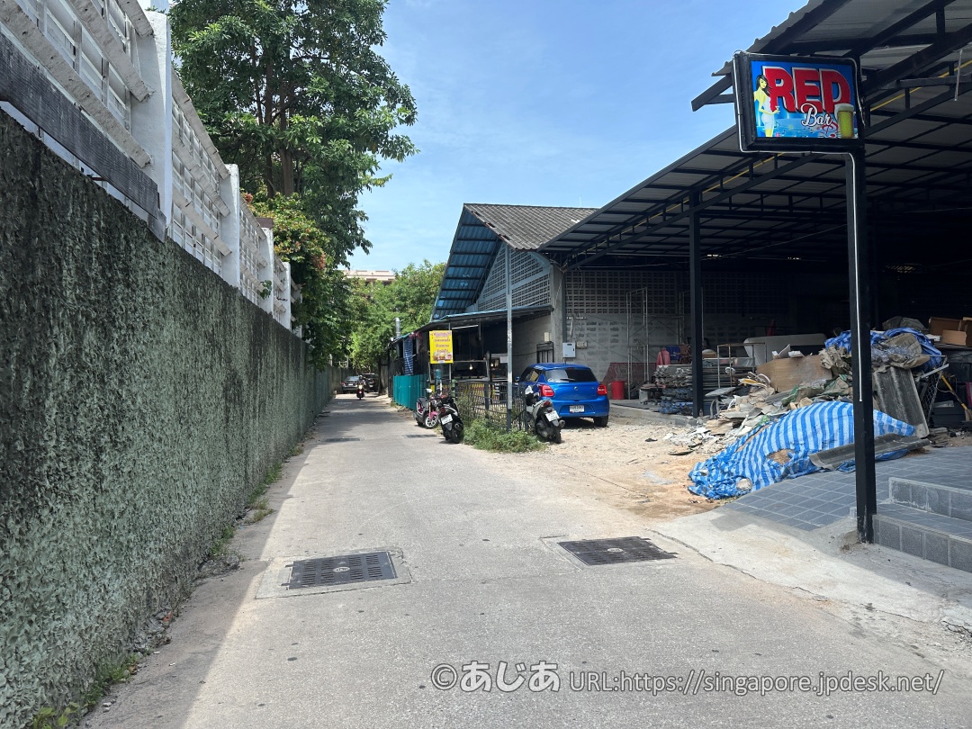コンクリートの塀に沿った細い路地。右側に「RED Bar」の看板があり、奥にトタン屋根の建物が見える。