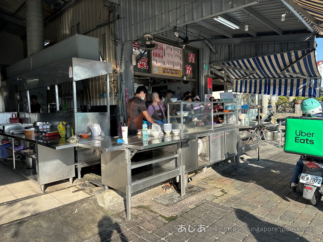 ステンレス製の調理台が並ぶ店頭で、料理を準備するスタッフと待機するUberEatsの配達員