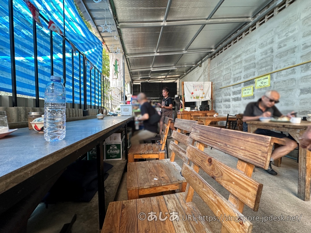 食堂の店内。木製のテーブルとベンチが並び、奥で黒いエプロンをしたスタッフが立っている。ローカルな雰囲気。