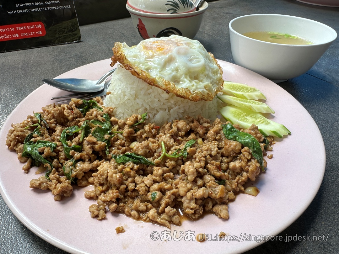 ピンク色のお皿に盛られたご飯、その上にカリカリの目玉焼き。手前にはたっぷりの豚ひき肉のバジル炒め(ガパオ)が添えられている。横にはスープの小鉢。