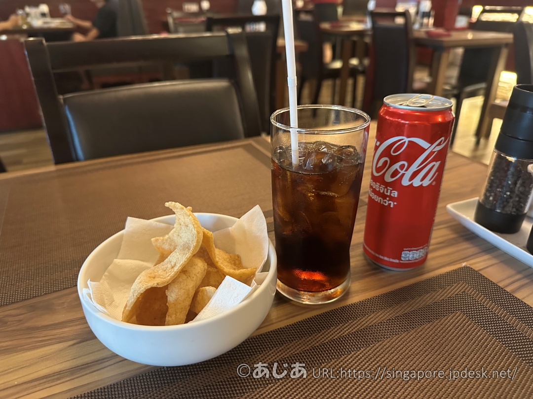テーブルに置かれた缶コーラ、氷入りのグラス、そして白いボウルに入った揚げスナック。