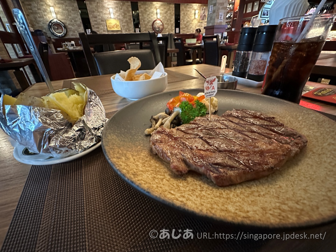 美しい模様の大きなお皿に乗ったリブアイステーキ。付け合わせの野菜と、別皿のベイクドポテト、奥にはコーラが見える。