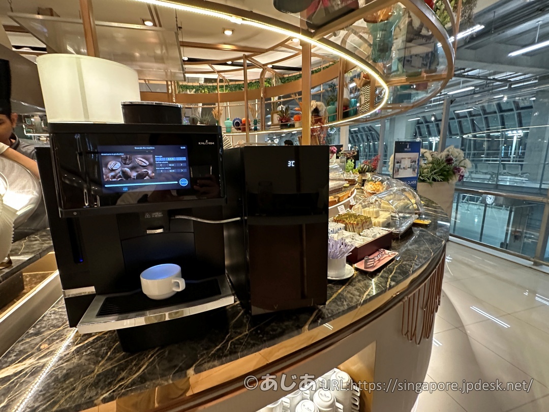 タッチパネル式の黒いコーヒーメーカーと、その横にあるガラスケース内のペストリーや軽食類。