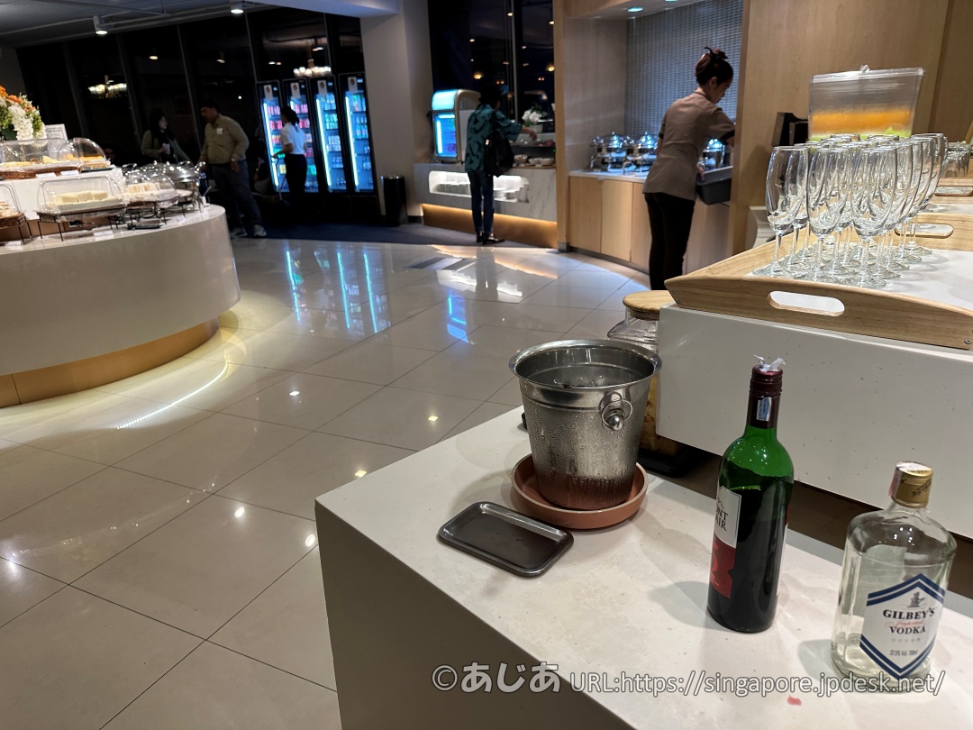 ビジネスクラスラウンジのダイニングエリア。手前にワインやウォッカの瓶があり、奥には料理の保温器や青く光るドリンク冷蔵庫が見える。