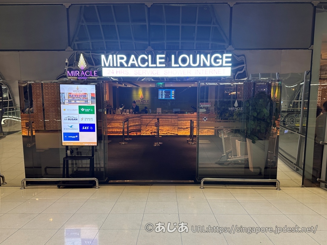 MIRACLE LOUNGEのエントランス正面。黒いカーペットとガラス扉があり、左側に提携航空会社やプライオリティパスのデジタル看板が立っている。