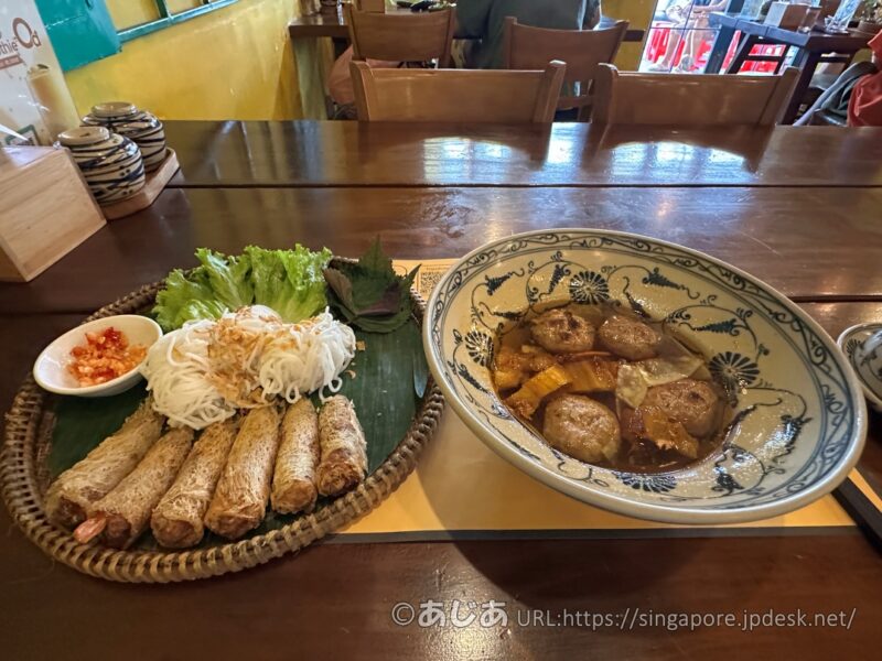 木製のテーブルの上に置かれた2つの料理。左は竹ザルに乗った揚げ春巻きとブン（米粉麺）、右はブルーの模様が入った陶器のどんぶりに入ったスープと肉団子。