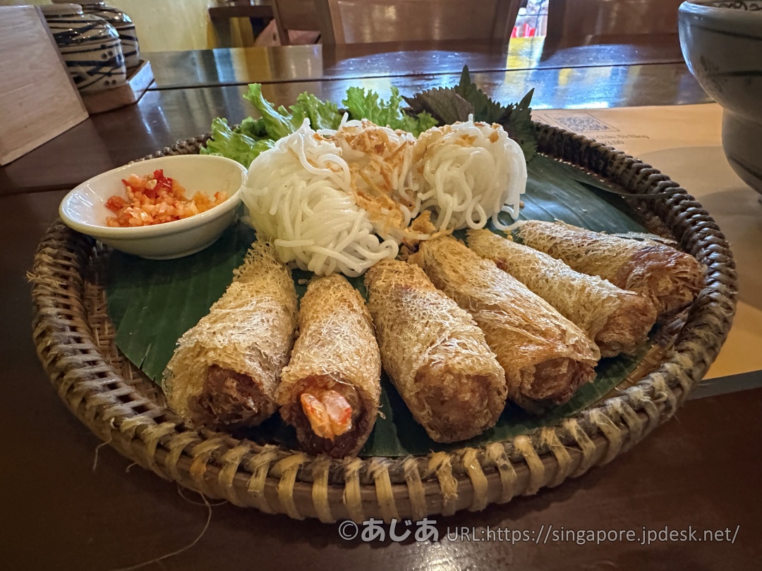 竹ザルに盛られた料理のアップ。手前には網目状のライスペーパーで巻かれた揚げ春巻きが並び、奥には白いブン（米粉麺）とレタス、シソなどのハーブ、左側にチリソースが添えられている。
