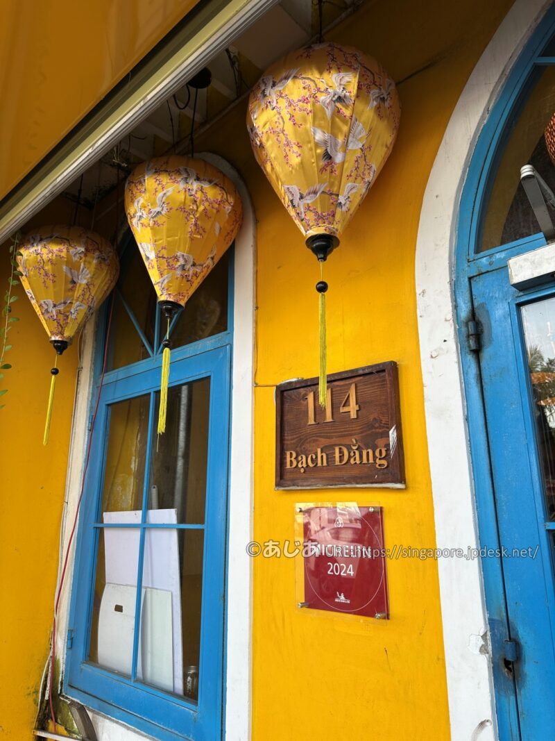 黄色い壁に飾られたホイアンランタンと、その下にある「114 Bạch Đằng」の住所プレート、「MICHELIN 2024」の赤いアクリルプレート。