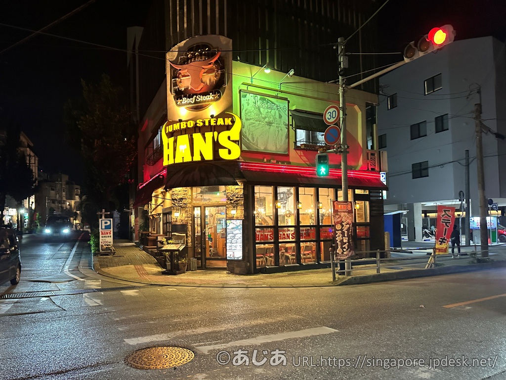 ジャンボステーキHAN'Sの外観。夜に黄色と赤のネオンサインが明るく光り、牛のマークが掲げられている角地の店舗。