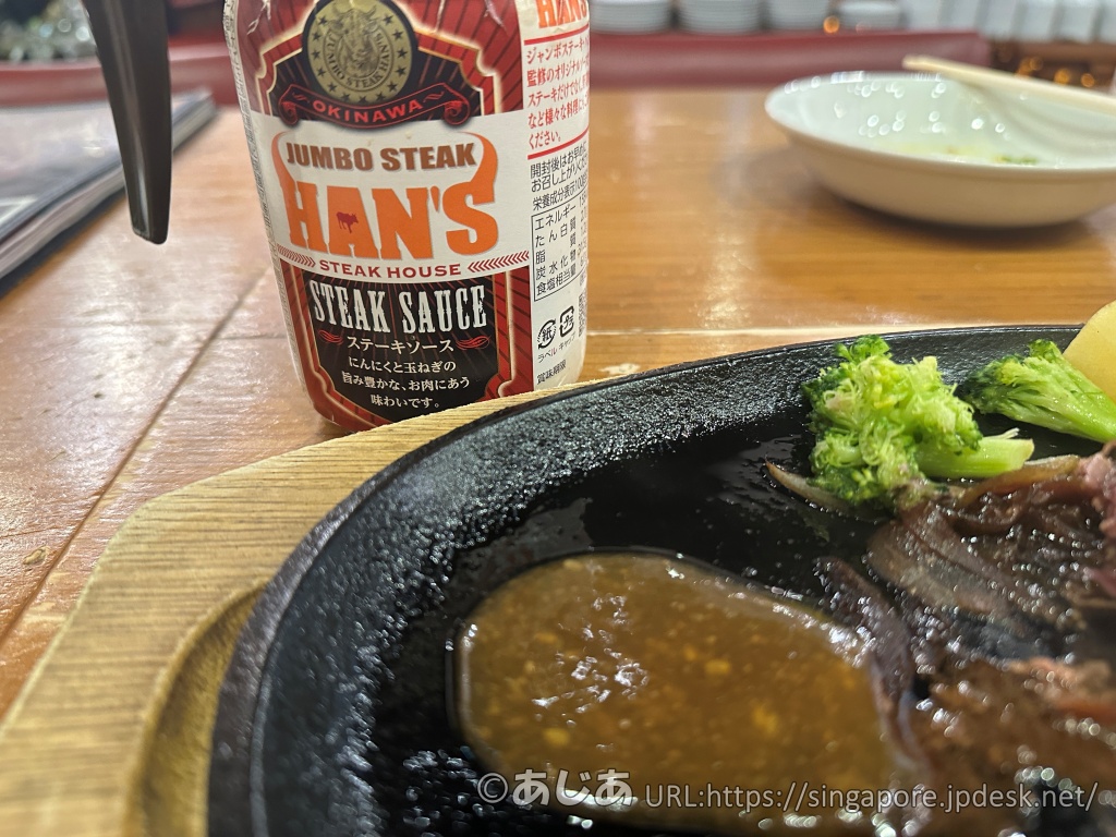 HAN'Sオリジナルステーキソースのボトル。手前の鉄板にはソースがたっぷりと注がれ、奥にはブロッコリーなどの付け合わせが見える。