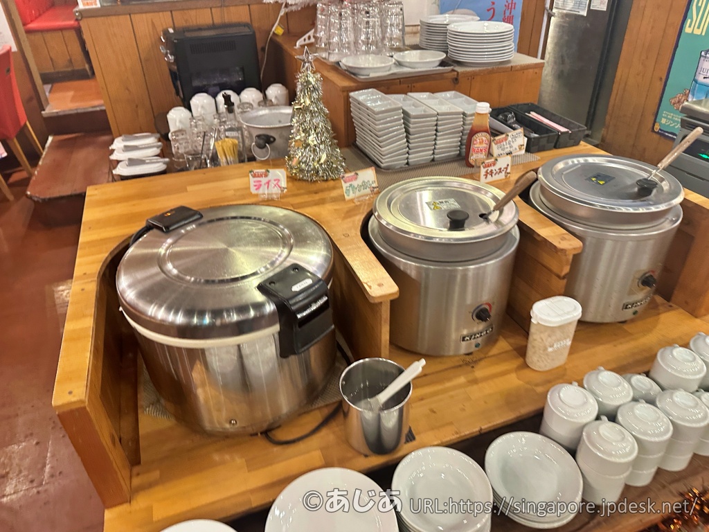 セルフエリアの様子。大きな炊飯器の隣にシルバーの保温ポットが2つ並んでおり、それぞれ「タイグリーンカレー」「チキンスープ」のポップが立てられている。