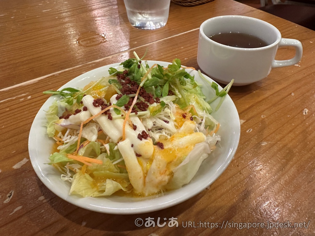 木製のテーブルの上に置かれたサラダとスープ。サラダには数種類の野菜に白いドレッシングとベーコンビッツがかかっている。奥にはマグカップに入ったスープがある。