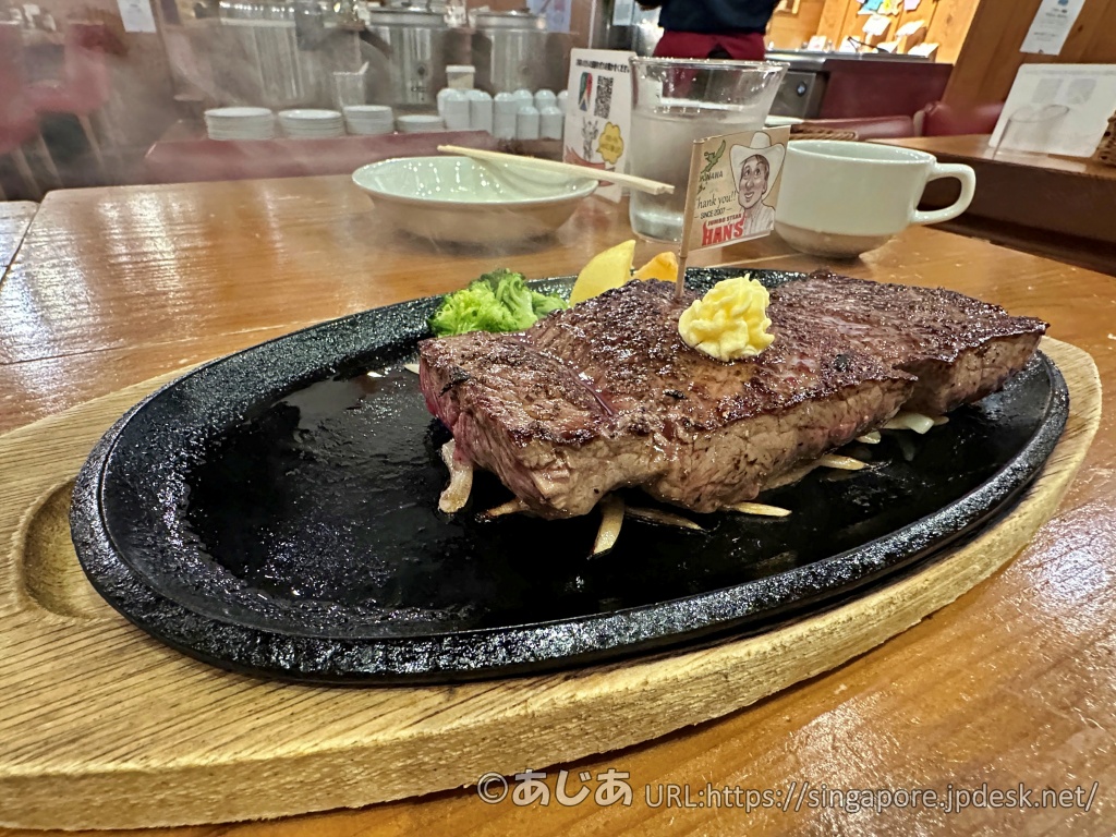黒い鉄板に乗った分厚いランプステーキ。上にはバターと、HAN'Sのキャラクターが描かれた小さな旗が刺さっている。鉄板からは熱々の湯気が立ち上っている。