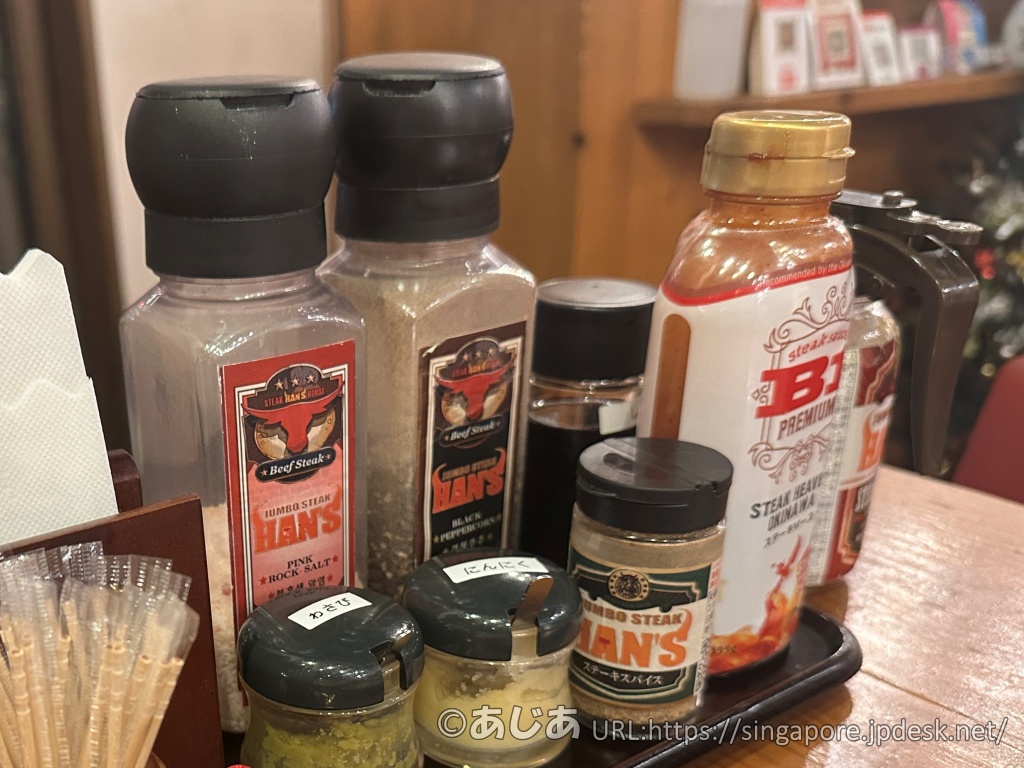 テーブルに並べられた調味料セット。ピンク岩塩、ブラックペッパー、わさび、にんにく、HAN'S特製ステーキスパイス、ステーキソースなどがカゴに入っている。