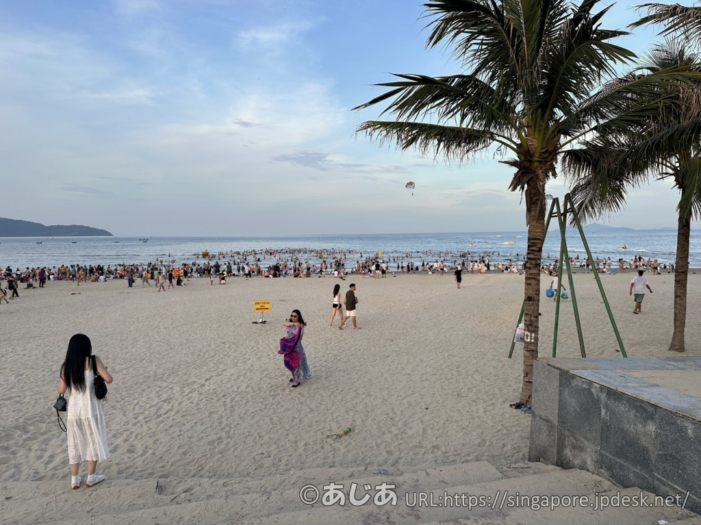 砂浜と海にたくさんの人が溢れ、活気づいている夕暮れ時のミーケービーチ。右手にはヤシの木が見える。